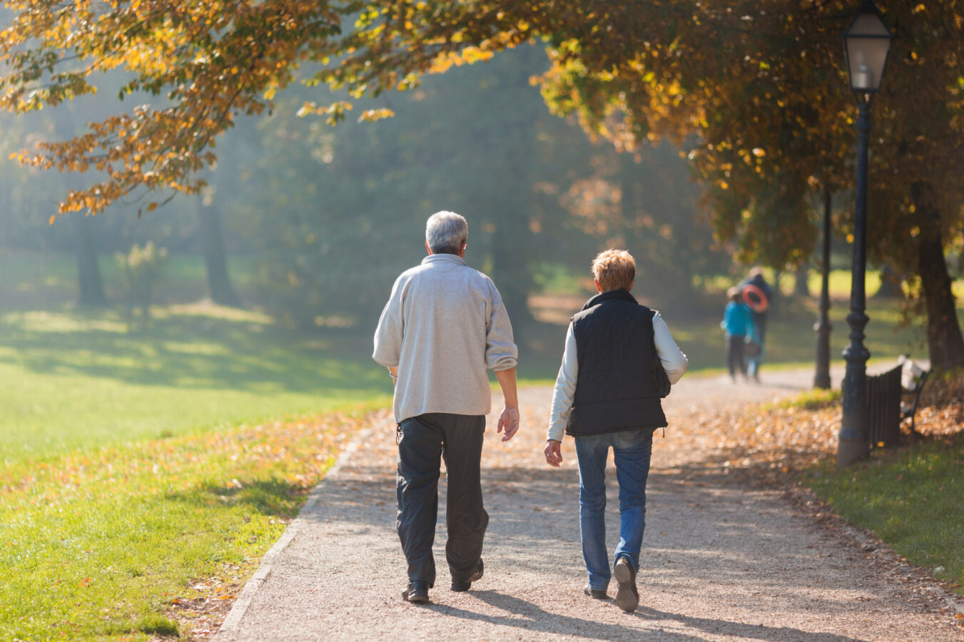 An elderly couple walks down a paved pathway in the fall, away from the camera.
