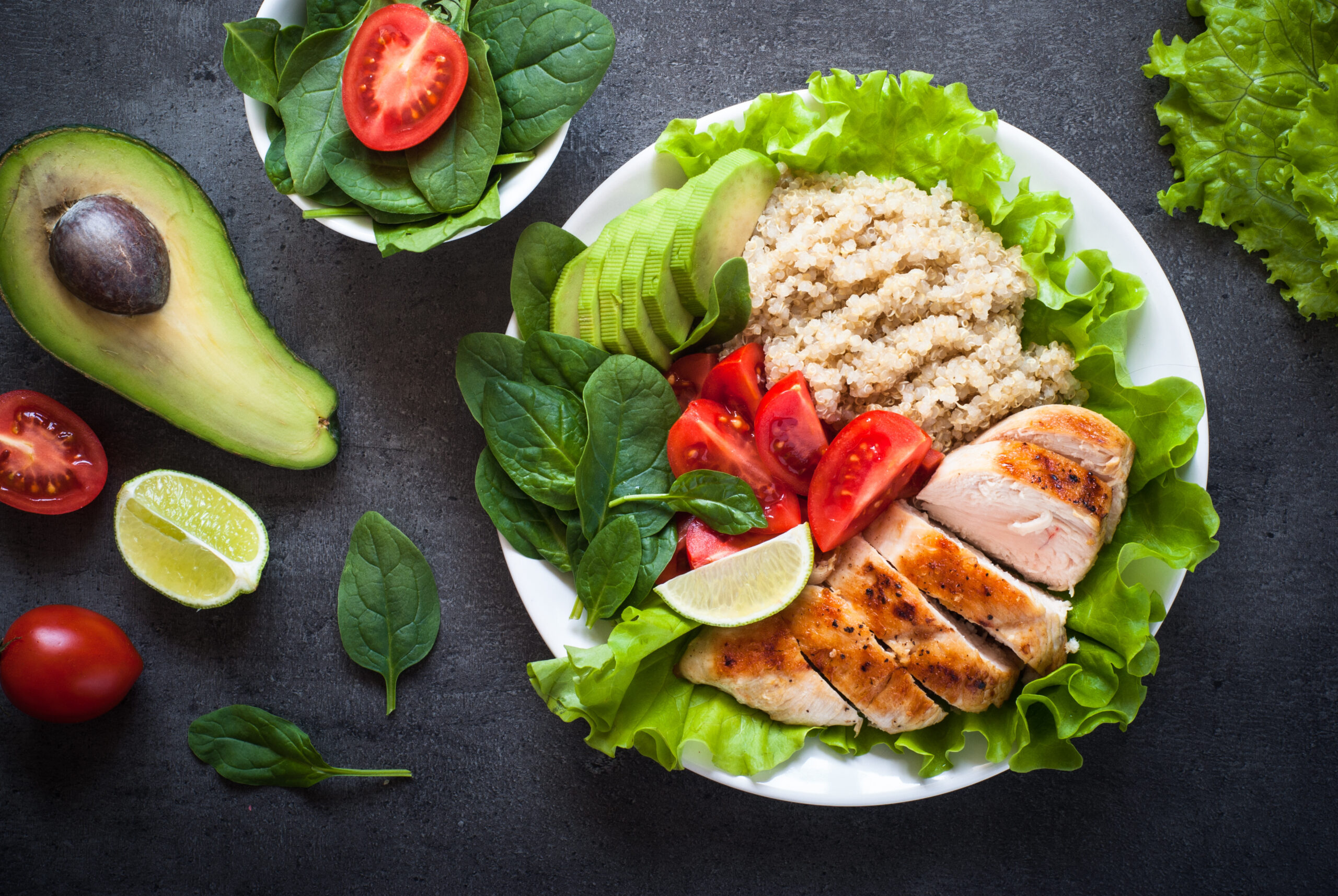 A salad is set on a white plate with chicken, tomatoes, quinoa and avocados on a dark table.