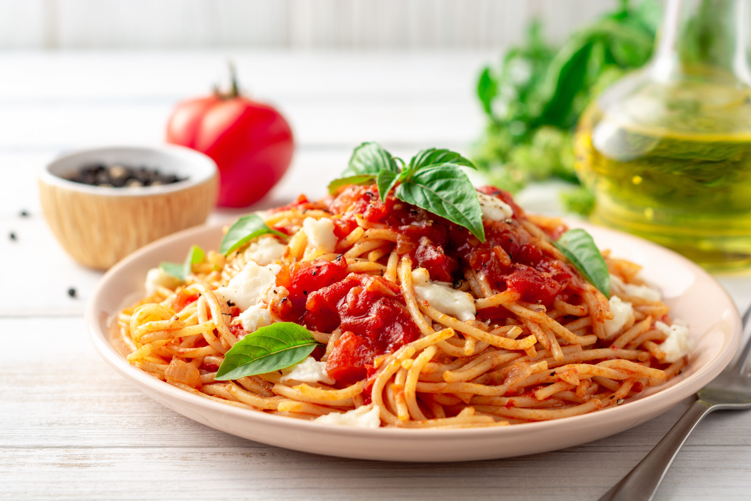 A plate of spaghetti sits on a table with pepper, tomato, and olive oil with basil on top.