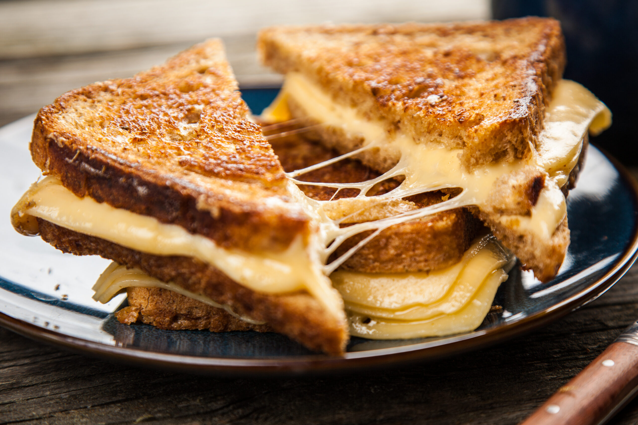 Two grilled cheese sandwiches sit on a dark plate, the top one is sliced, demonstrating a delicious cheese-pull.