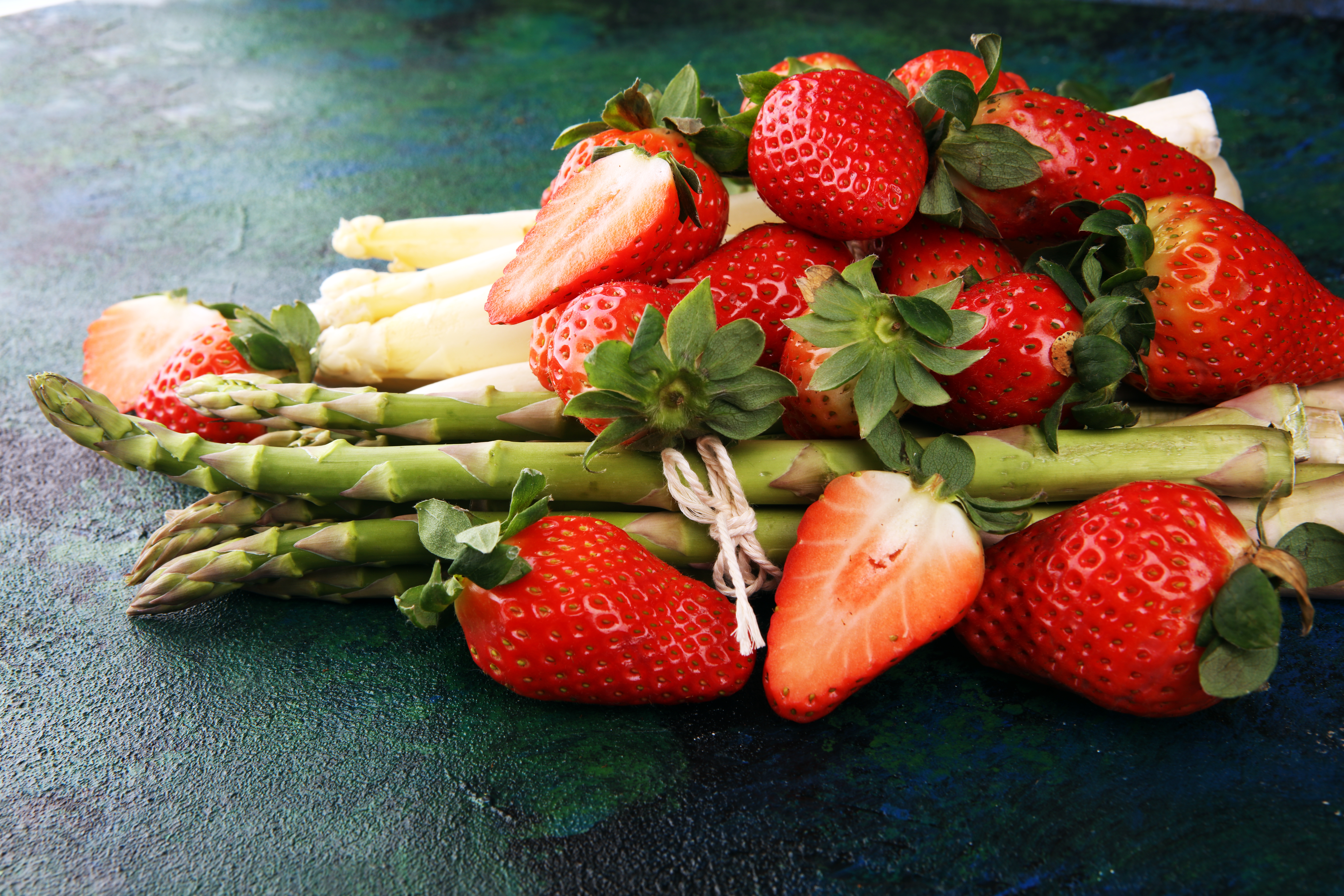 Whole and sliced strawberries laying over asparagus on a black table.