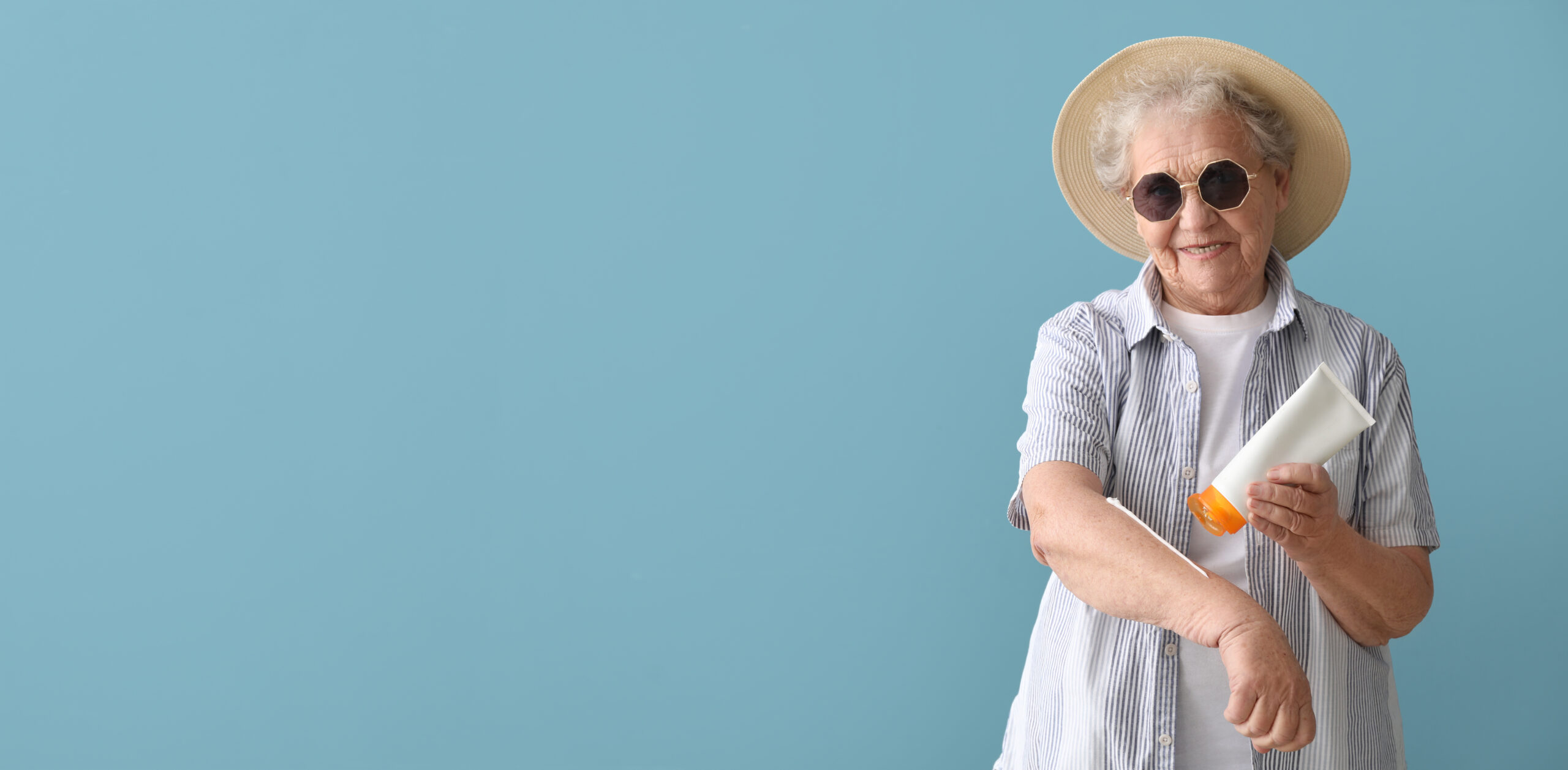 An elderly woman in a sun hat and sunglasses applies sunscreen to her arm in front of a blue background.