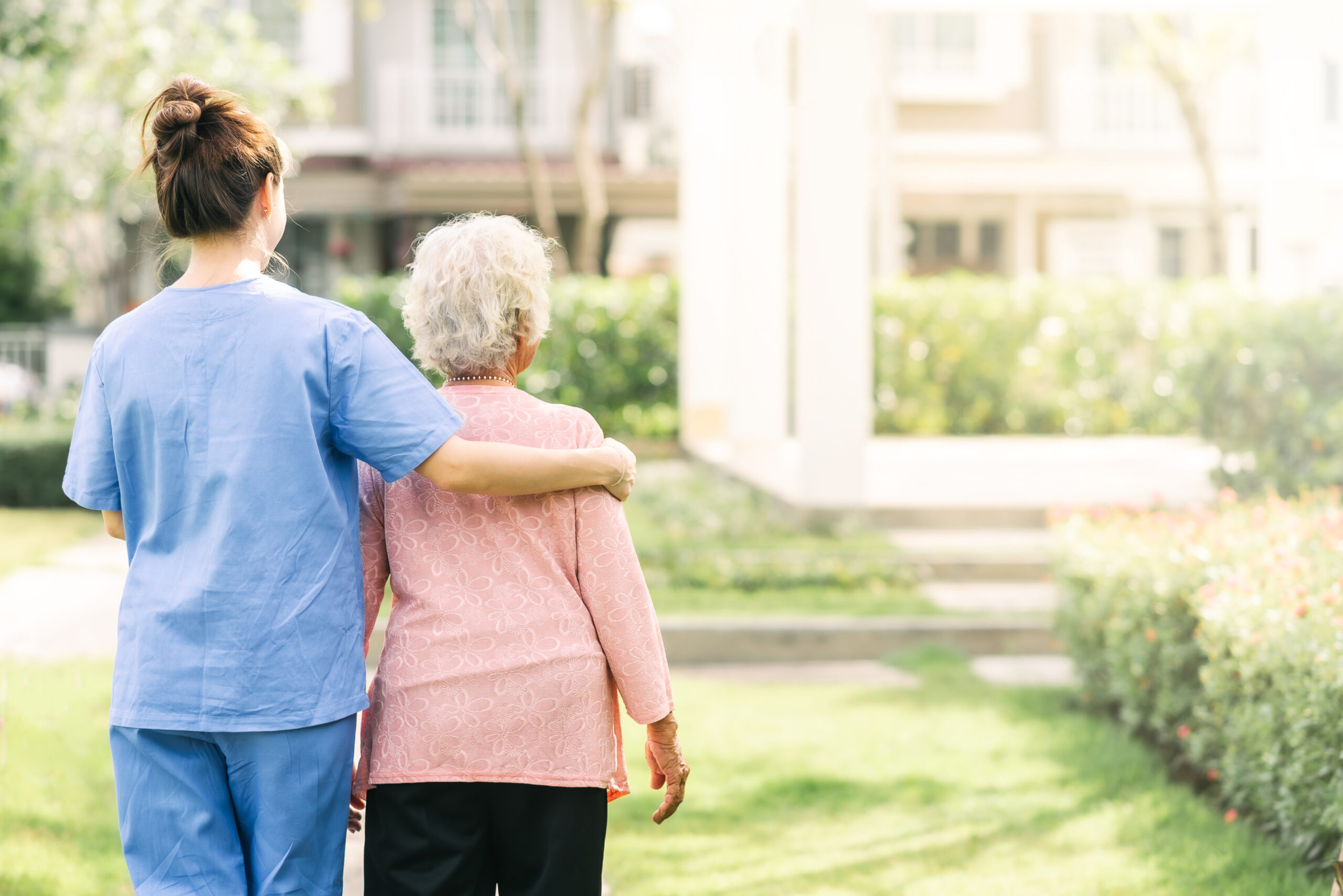 A caregiver walks with an elderly woman outside, surrounded by greenery and sunshine.