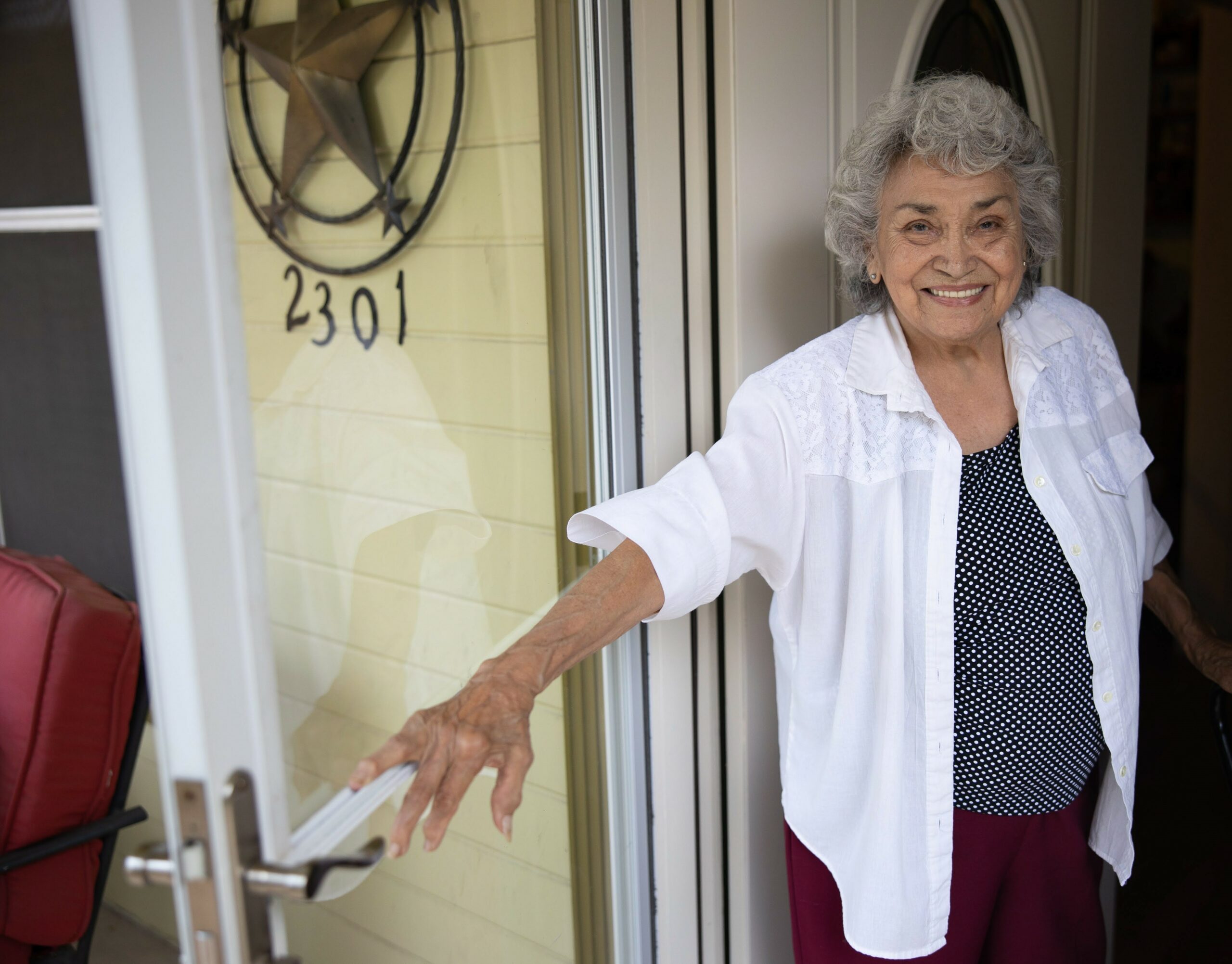 An elderly women opening her storm door to the camera with a smile as she receives home delivered meals.