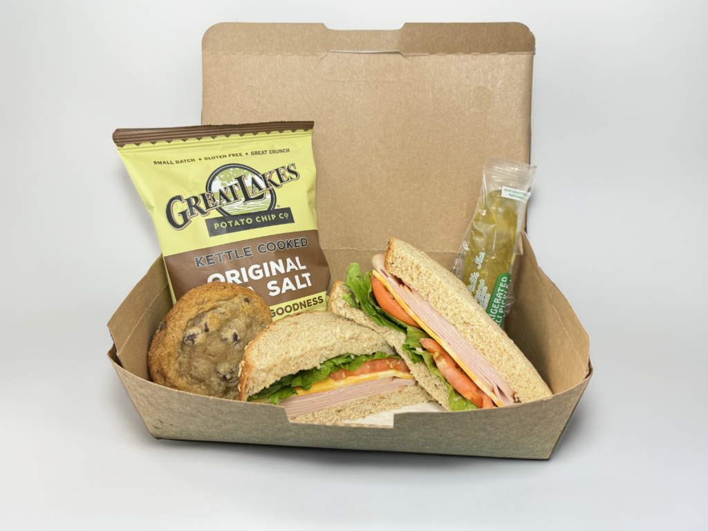 A sandwich, cookie, bag of chips, and a pickle in a More Than a Meal Catering box.