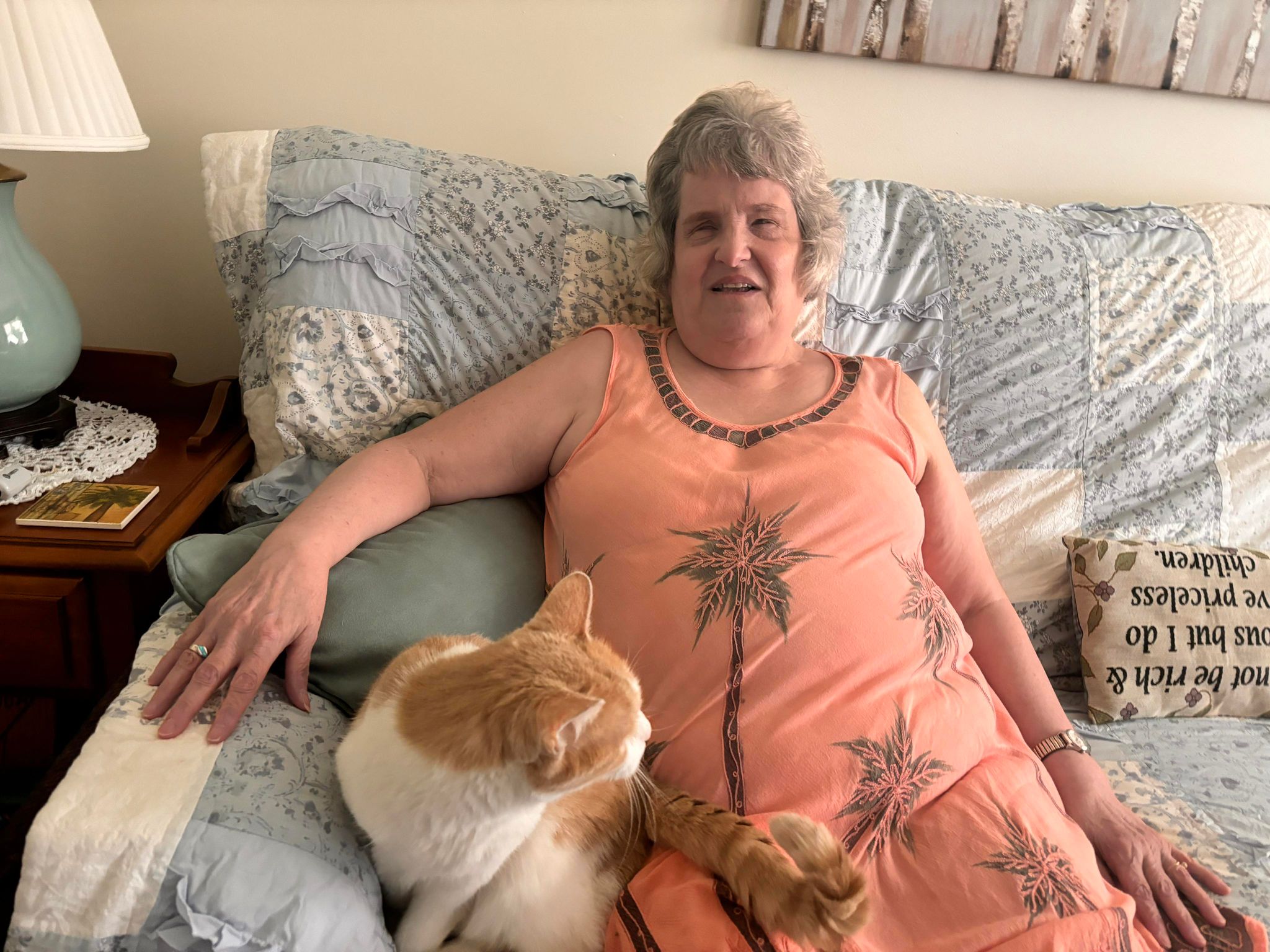 Linda sits in her home on the couch holding her cat Jordie while her home delivered meals program driver helps her put her meals away.